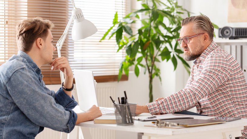 Ein älterer Mann am Schreibtisch beträt einen jüngeren Mann